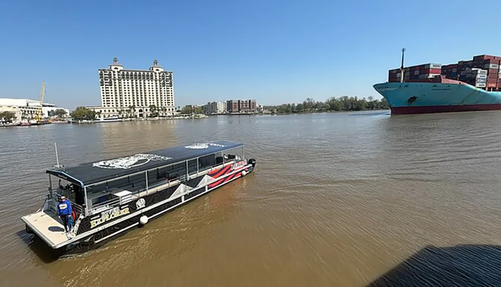 A riverboat cruises on a wide river with a cargo ship in the background near urban buildings under a clear blue sky