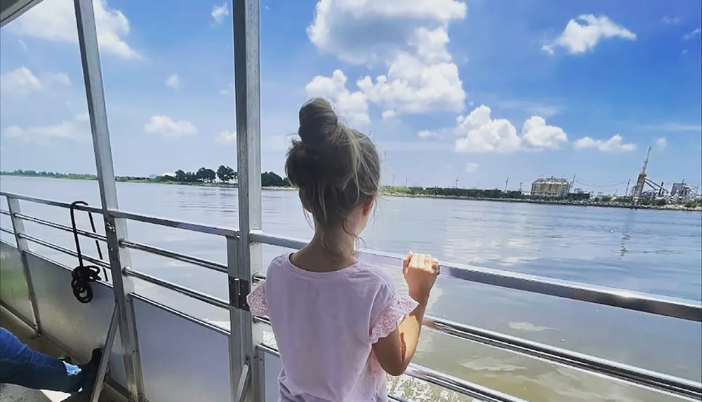 A child gazes out over the river from the deck of a boat with industrial structures visible on the distant shore under a partly cloudy sky