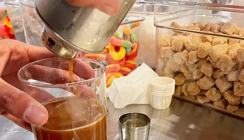 A person is pouring a dark liquid into a plastic cup likely a beverage with a background featuring containers of colorful cereal