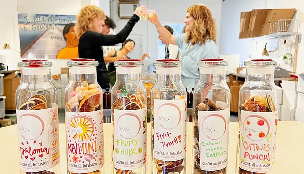 Foreground features a set of cocktail infusion bottles while in the background two people are happily toasting with drinks in a lively friendly atmosphere