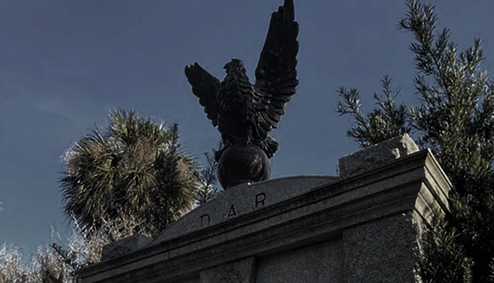 The image shows a dark dramatic statue of an eagle with spread wings perched atop a pedestal with the letters DAR inscribed on it surrounded by a clear sky and foliage