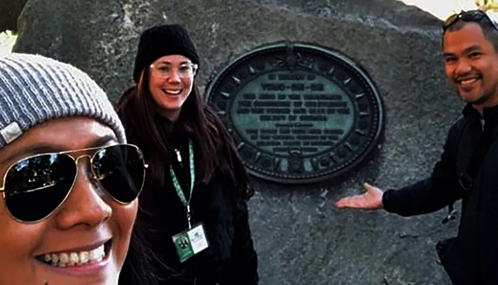 Three people are posing for a selfie with one of them pointing to a commemorative plaque