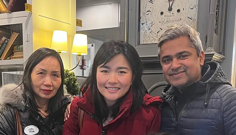 Three people are smiling for the camera in a cozy interior with decorative items and a clock in the background