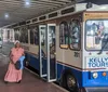 The image shows a trolley-style tour bus marked Kelly Tours with passengers on board stopped under a covered area where pedestrians are walking with a zoomed-in inset of a dish that appears to be seasoned shrimp