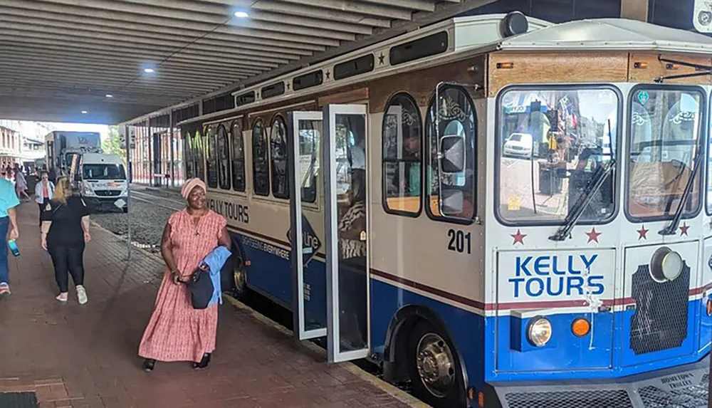 A woman is smiling near a Kelly Tours trolley with other pedestrians nearby under a sheltered walkway