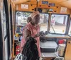 The image shows a trolley-style tour bus marked Kelly Tours with passengers on board stopped under a covered area where pedestrians are walking with a zoomed-in inset of a dish that appears to be seasoned shrimp