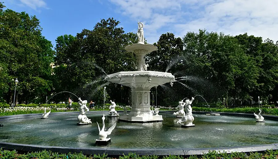 An ornate white fountain featuring statues and multiple water jets is set amidst a lush green park under a blue sky.
