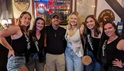 A group of five women wearing sashes with bridal party titles and a man posing together at a bar, possibly celebrating a bachelorette party.