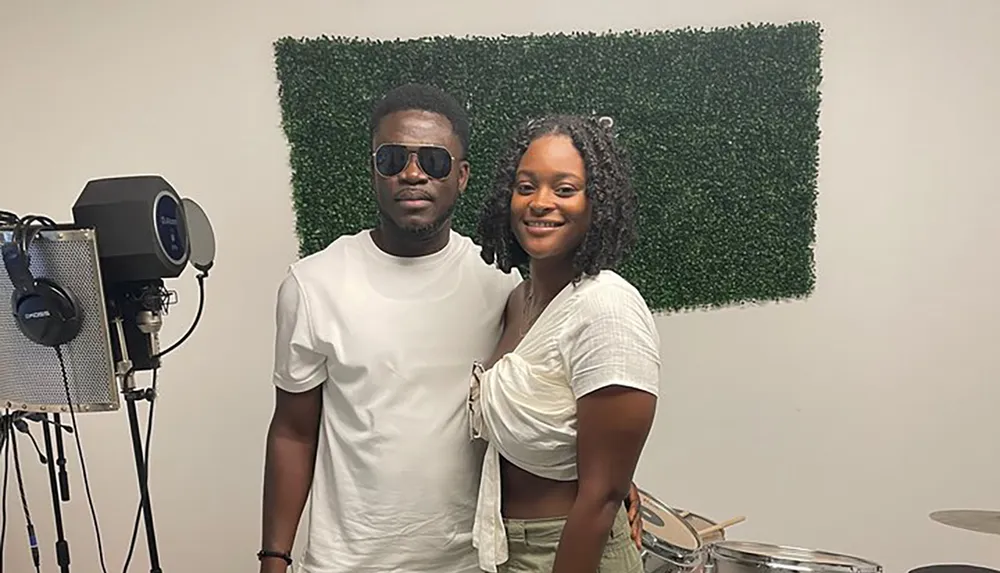 A man and a woman are smiling for a photo in a room with music equipment and a wall decorated with artificial greenery