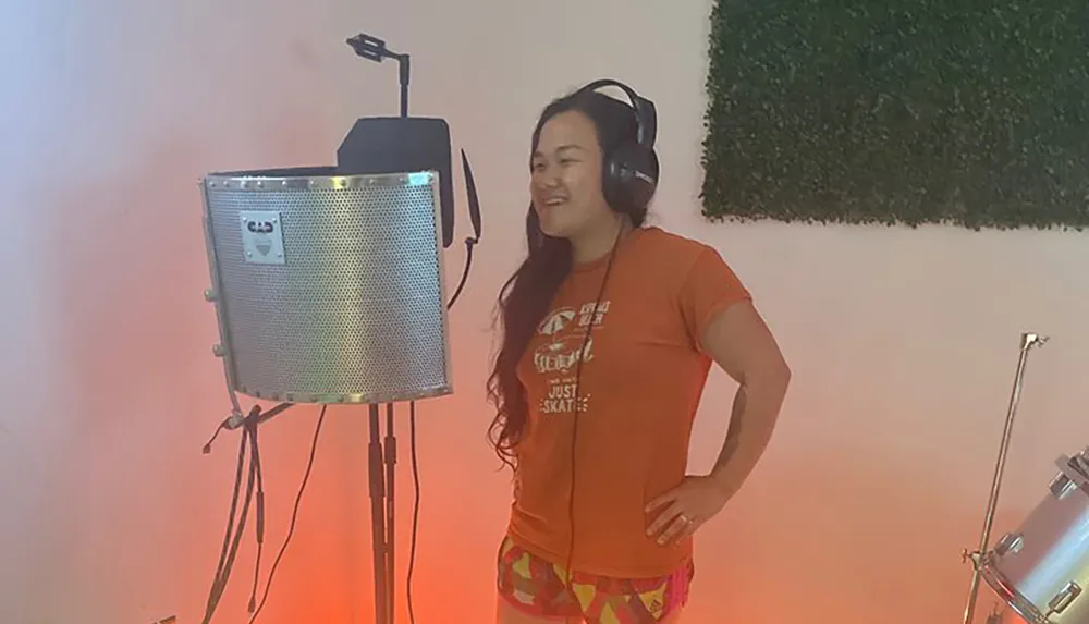 A person with headphones stands smiling in a recording studio in front of a microphone with a pop filter