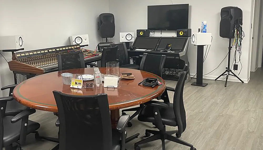 The image shows a professional audio mixing and recording studio setup with a large analog mixing console speakers acoustic treatment and a round meeting table with chairs