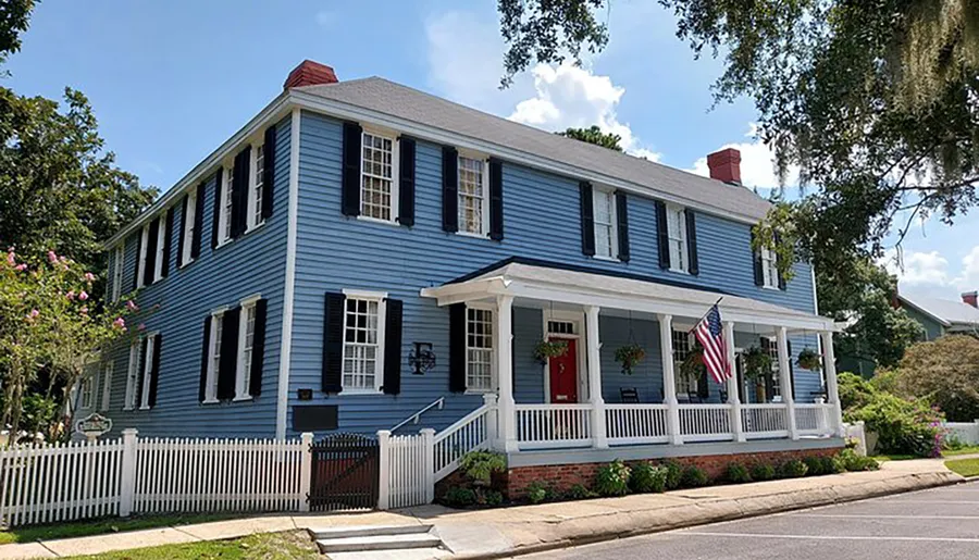 A traditional two-story blue house with white shutters, a red door, and an American flag displayed on the front porch, surrounded by a white picket fence.