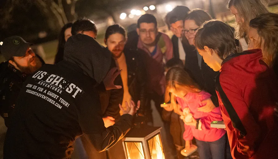 A group of people attentively gathers around a lantern held by a guide wearing a hood and a US Ghost Adventures jacket during what appears to be a ghost tour at night.