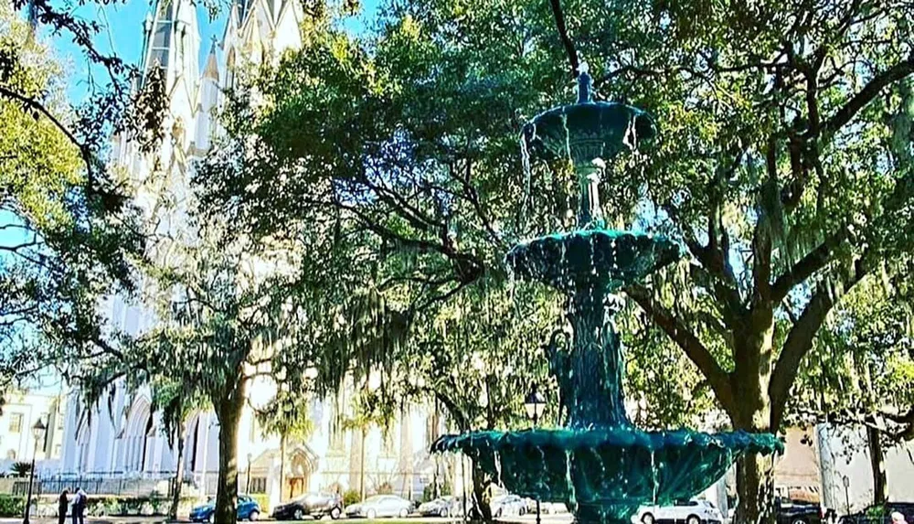 A verdigris-colored tiered fountain stands in front of a cathedral with gothic architectural features surrounded by trees draped in Spanish moss under a clear blue sky