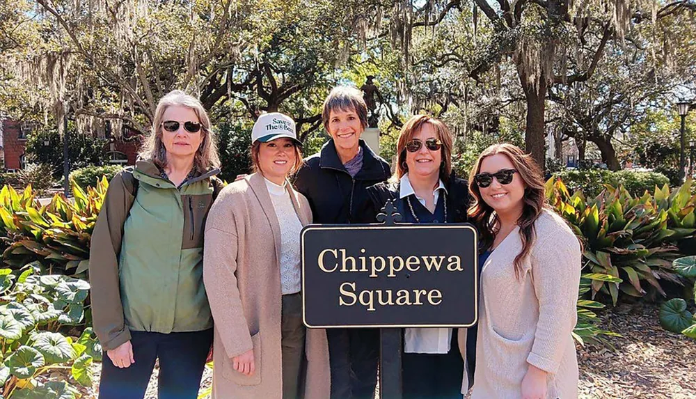 Five women are posing for a group photo in front of the Chippewa Square sign with trees and foliage in the background