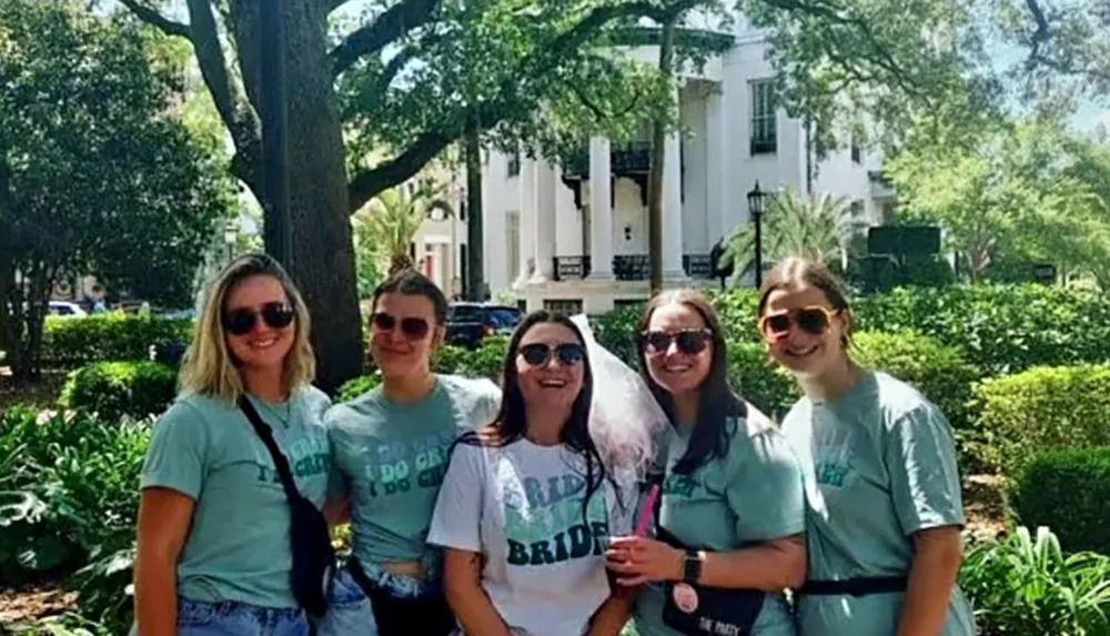 A group of five individuals presumably on a bachelorette party as indicated by their matching shirts and the bridal veil are posing for a photo in front of a classic white building with large columns