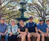 Six people are sitting in front of a green multi-tiered fountain with a backdrop of lush trees on a sunny day