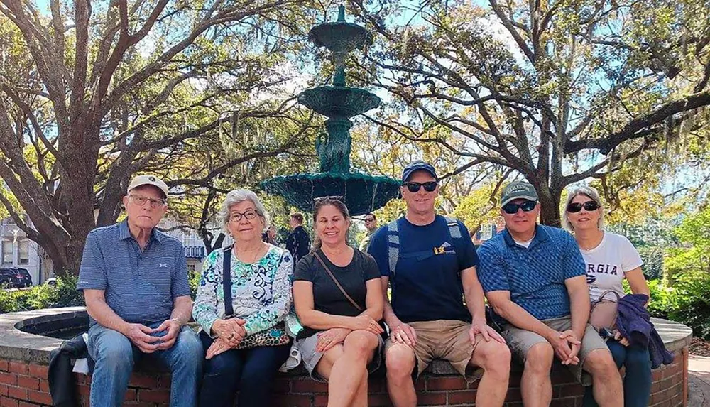 Six people are sitting in front of a green multi-tiered fountain with a backdrop of lush trees on a sunny day