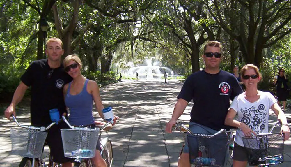 Four individuals are posing for a photo with their bicycles on a tree-lined path with a fountain visible in the background