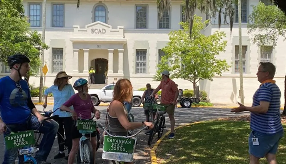 A group of people on a bike tour are listening to a guide in front of a building marked SCAD