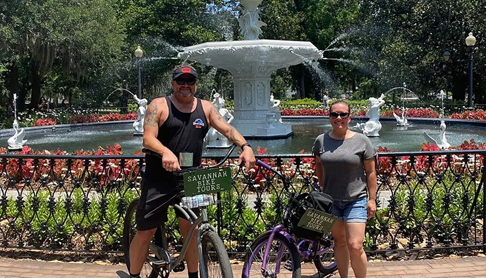 Two people with bicycles are standing in front of a fountain in a sunny park setting likely enjoying a day of sightseeing