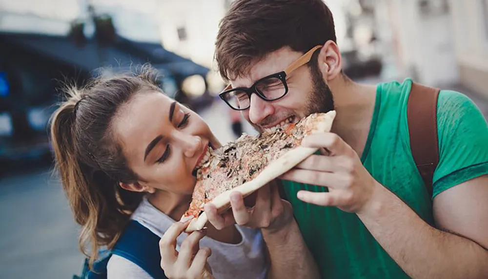 A man and a woman are joyfully sharing a large slice of pizza on a sunny day