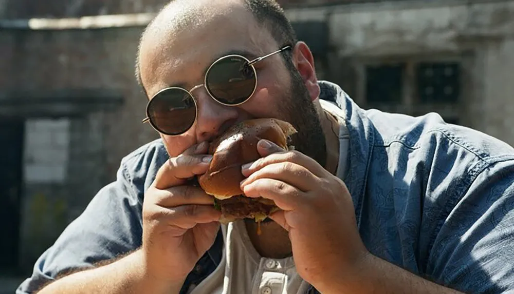 A man with a beard and round sunglasses is taking a big bite of a hamburger