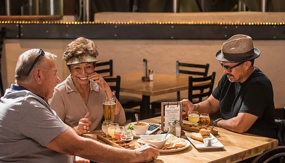 Three people are sharing a meal and drinks at an outdoor restaurant with one person smiling and wiping away a tear another person listening attentively and a third person focused on something at the table