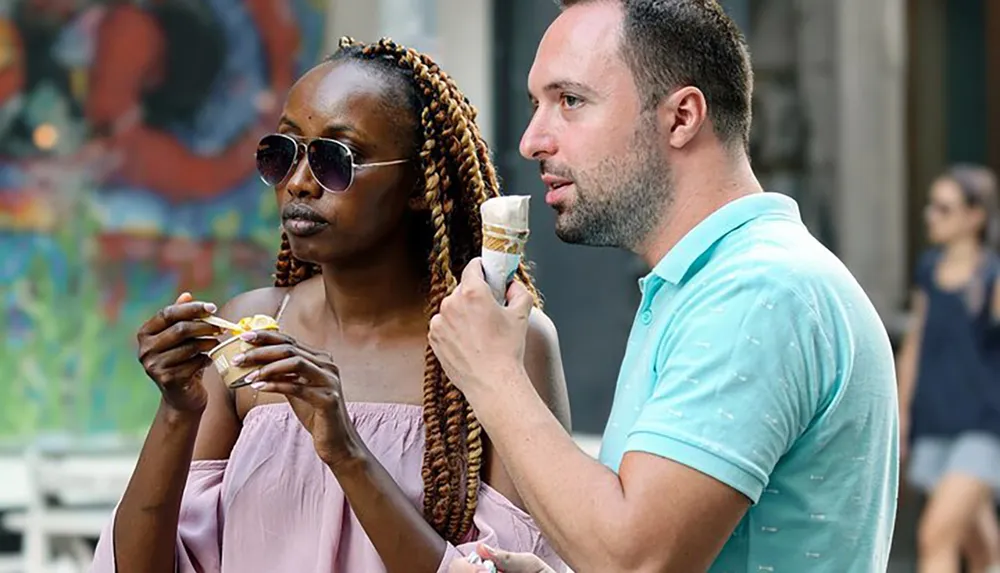 A woman and a man are standing outside as they enjoy eating ice cream on a sunny day