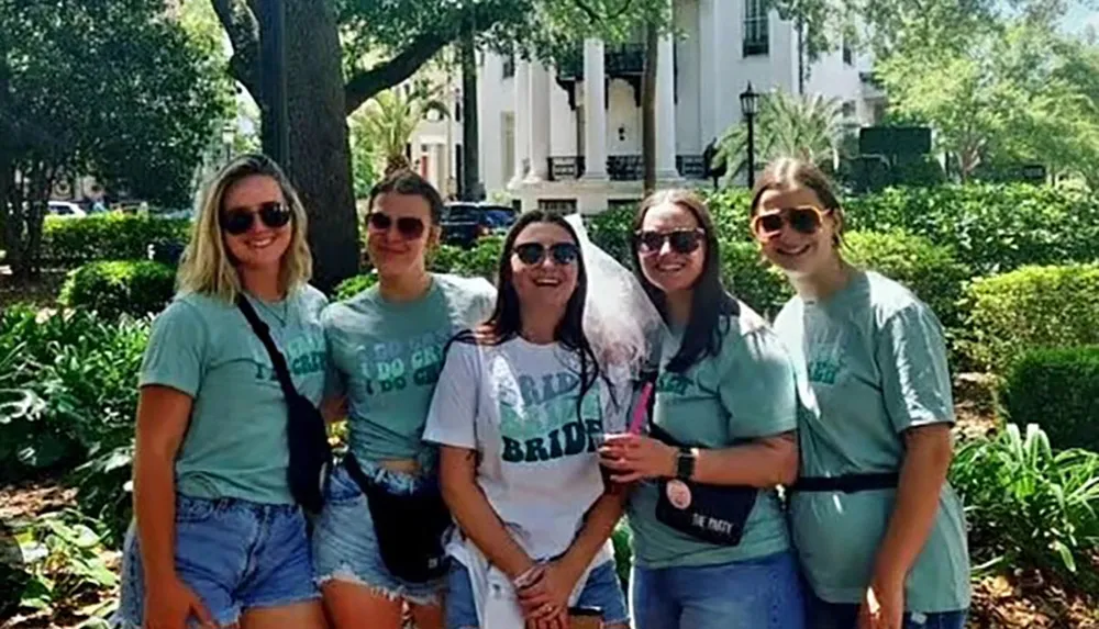 A group of five women wearing matching t-shirts are smiling for the camera with one wearing a veil suggesting a bridal party event