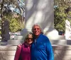 Two people are smiling for a photo in front of a monument flanked by lion statues in a park with trees in the background