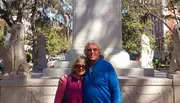 Two people are smiling for a photo in front of a monument flanked by lion statues in a park with trees in the background.