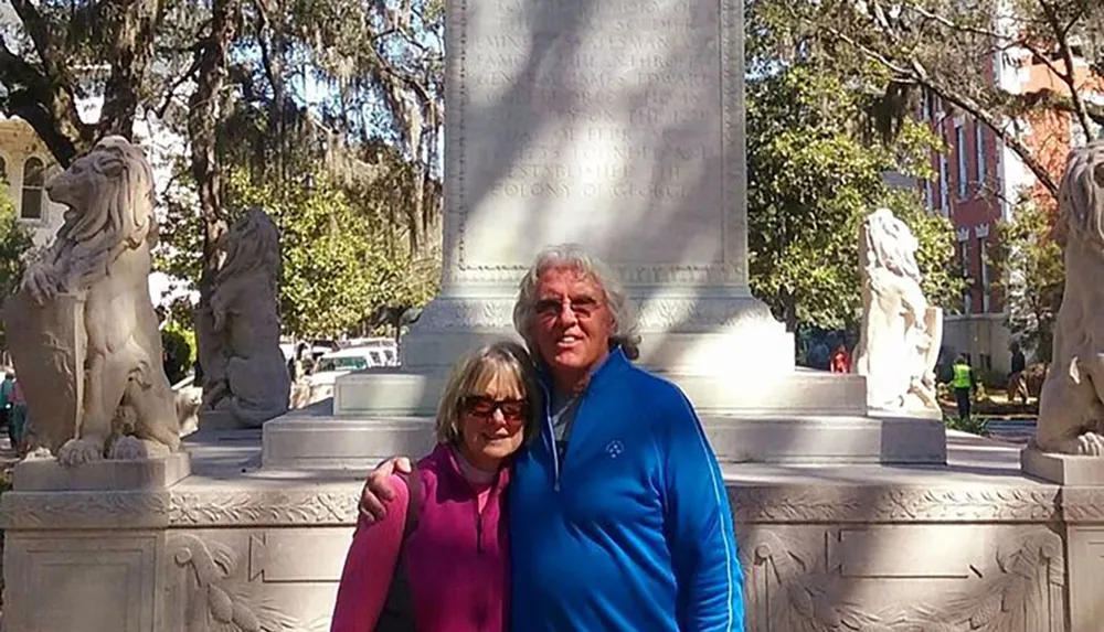 Two people are smiling for a photo in front of a monument flanked by lion statues in a park with trees in the background