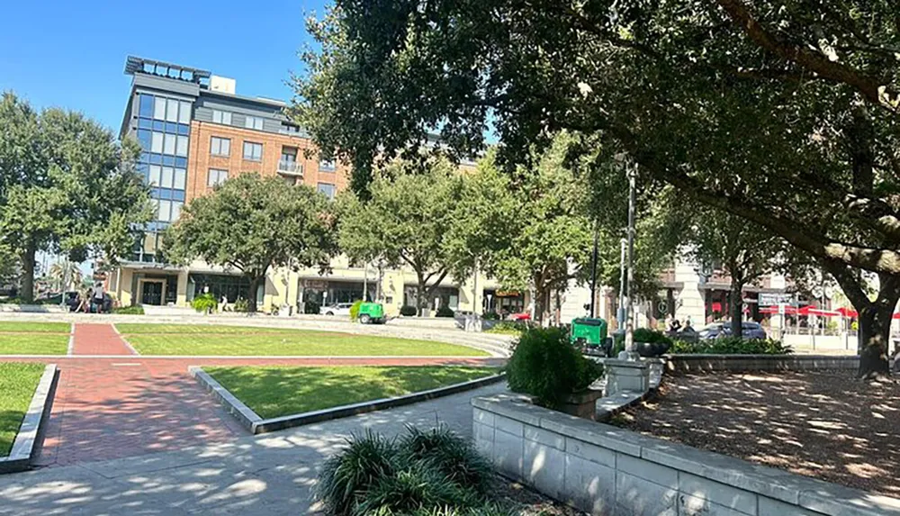 The image shows a sunny day in an urban park with a well-manicured lawn walking paths mature trees street furniture and a city building in the background