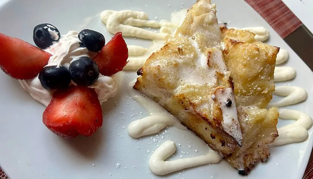 A slice of bread pudding is garnished with whipped cream fresh strawberries blueberries and a decorative drizzle of sauce on a white plate