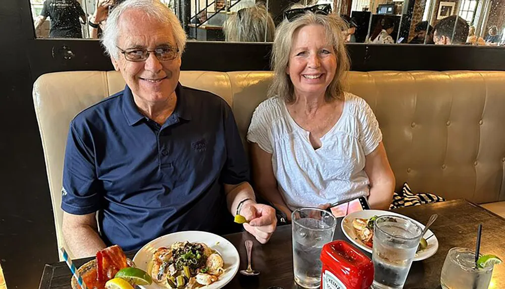 A smiling man and woman are sitting in a booth at a restaurant with plates of food in front of them and drinks on the table