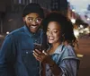 A smiling man and woman are sharing earphones and looking at a smartphone together on a city street at dusk