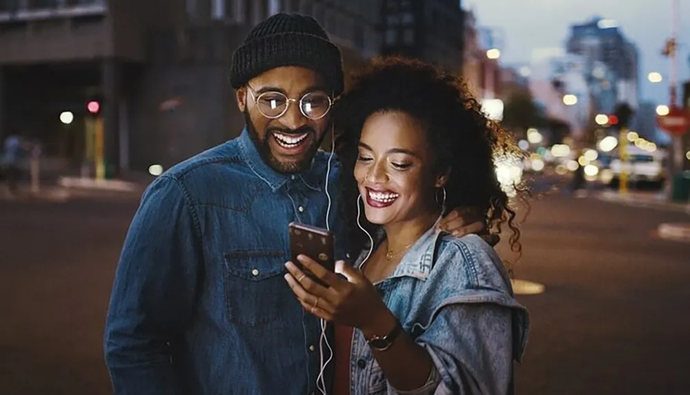 A smiling man and woman are sharing earphones and looking at a smartphone together on a city street at dusk