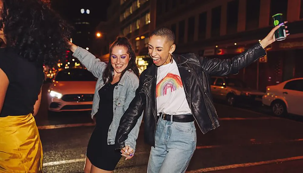 Two joyful people are holding hands and dancing on a city street at night with one of them holding a drink can aloft