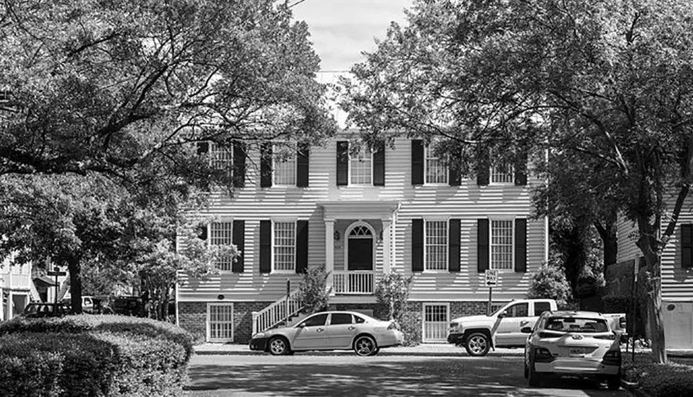 The image shows a two-story residential building with traditional architecture surrounded by trees with parked cars in front in a black and white photograph