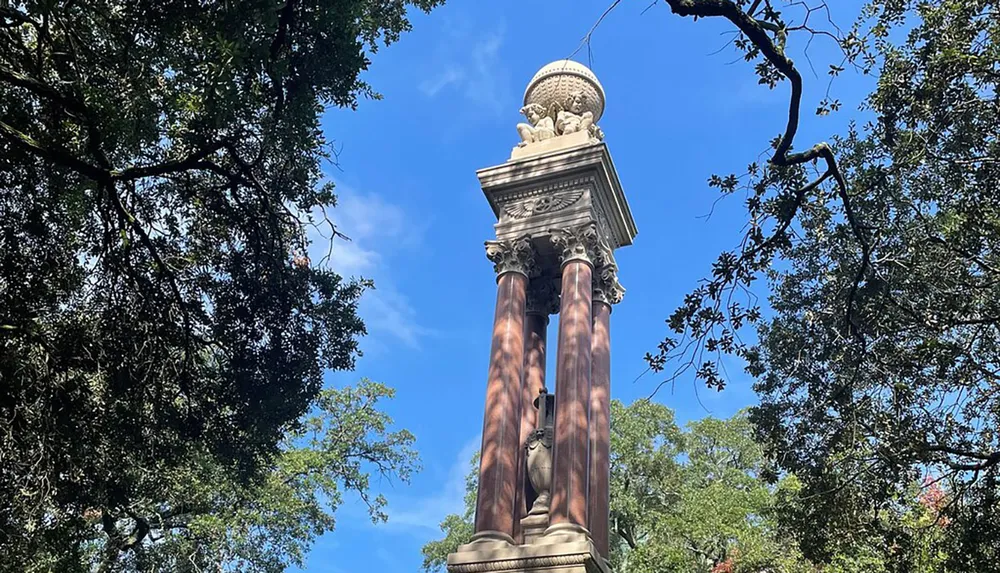 The image shows a tall ornate column with decorative elements set against a clear blue sky with lush green trees partially framing the view