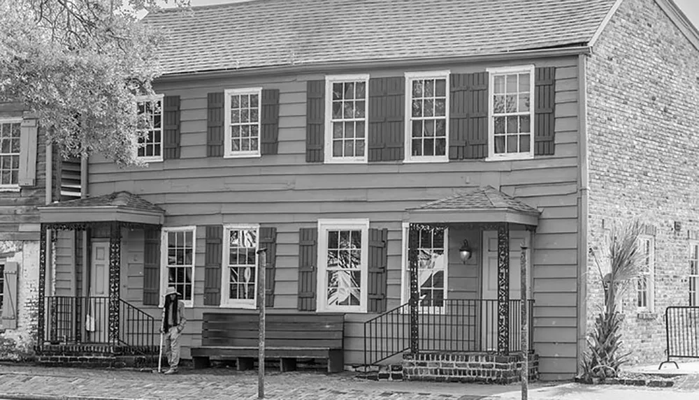 This black and white image depicts a historical two-story wooden building with shutters on the windows complete with a brick side facade and a person standing on the sidewalk in front