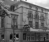 The black and white image features a classic theater facade with a marquee sign under a tree-lined street