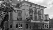 The black and white image features a classic theater facade with a marquee sign under a tree-lined street.