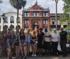 A diverse group of people is posing for a photo in front of a historic-looking building with a distinctive red facade and a lion statue on the rooftop