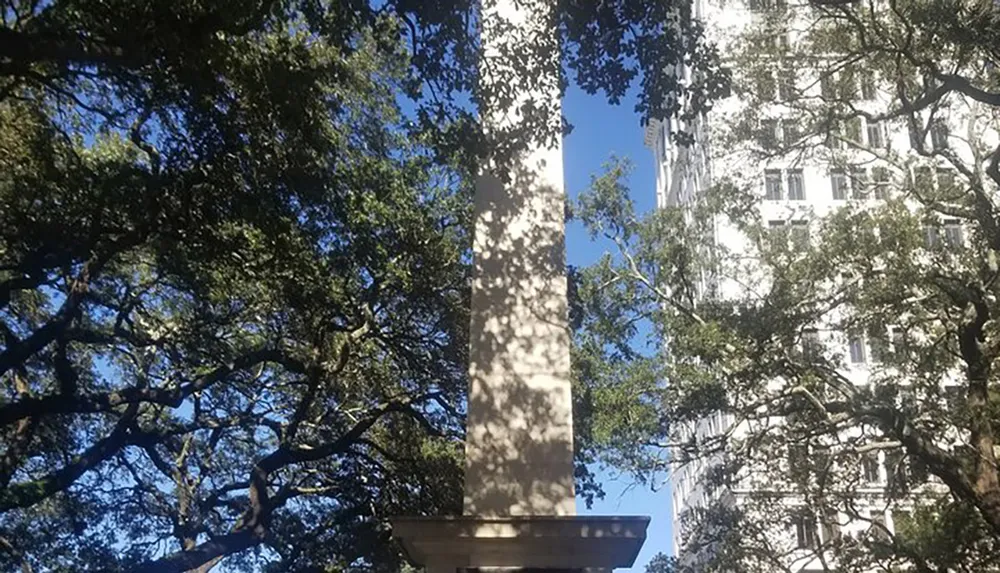 The image shows the base of a column or monument with lush green trees and a multi-story building in the background all under a clear blue sky