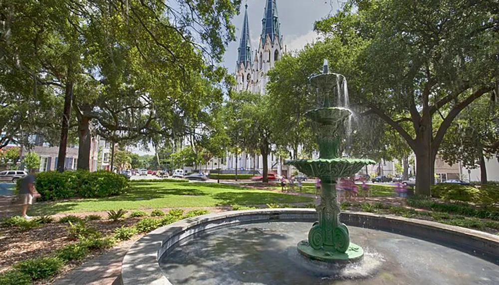 The image shows a verdant park with a green fountain in the foreground and an impressive Gothic-style cathedral in the background under a sunny sky with lush trees surrounding the scene