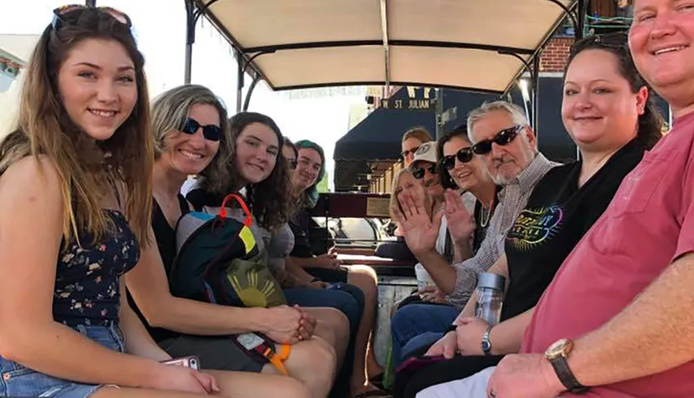 A diverse group of people are smiling and waving at the camera while seated on some sort of open-air group transport or tour vehicle