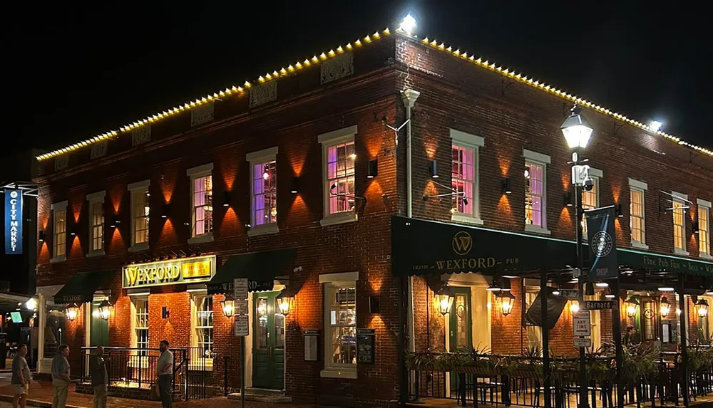 The image features a well-lit two-story brick building at night that houses Wexford Pub with people walking by on the sidewalk and festive lights strung along the roofline