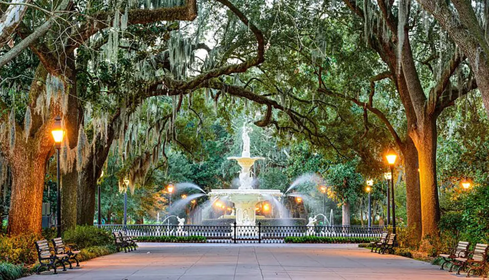 A picturesque fountain is illuminated at twilight surrounded by lush trees draped with Spanish moss in a serene park setting